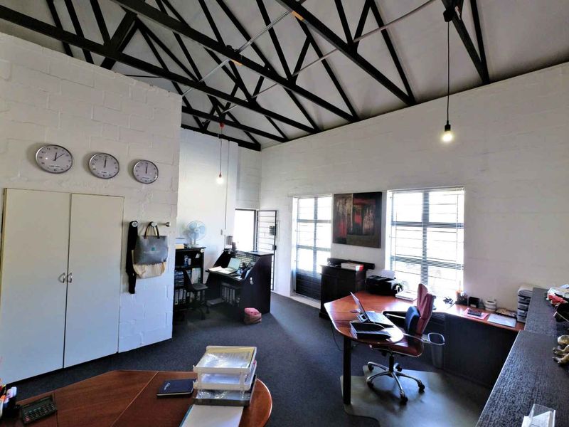 Spacious industrial-style office featuring exposed steel roof trusses, painted brick walls, and large windows with security bars, offering a functional workspace.