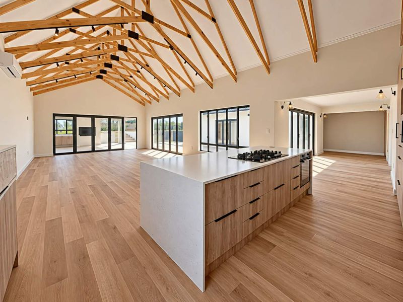 Spacious open-plan living area featuring high vaulted ceilings with exposed timber trusses, modern kitchen island with integrated appliances, and large glass sliding doors.
