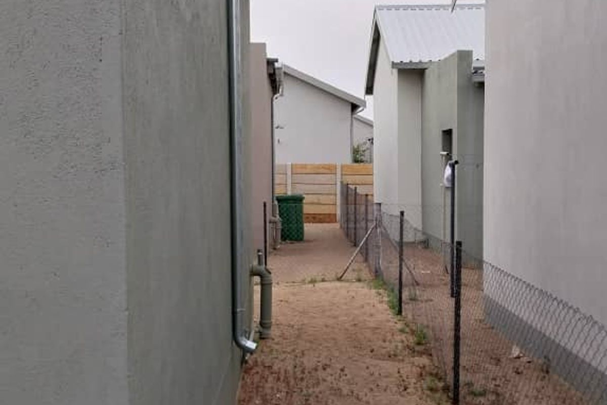 A narrow side passage between two residential structures featuring a metal downpipe, chain-link fencing, and an unpaved ground area.