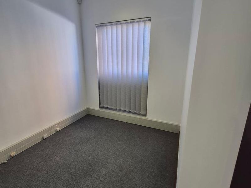 A compact, empty office space featuring grey carpeting, vertical window blinds, and a ceiling-mounted fluorescent light fixture.