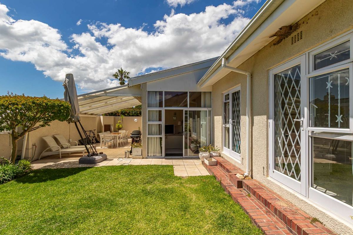 Rear exterior view featuring a paved covered patio, brick step transition, and sliding glass doors leading into the home, set against a landscaped lawn.