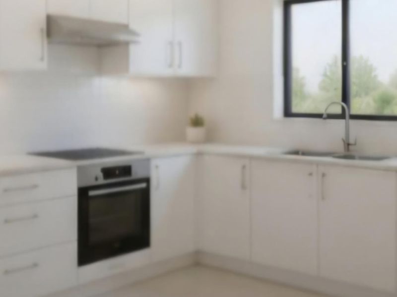 A modern, minimalist kitchen featuring white cabinetry, stone countertops, integrated oven, electric cooktop, and a double bowl sink with a view through the window.