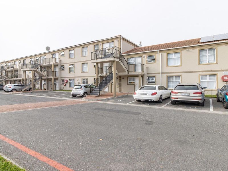 Exterior view of a multi-story apartment complex featuring designated parking bays, external staircases, and a tiled roof with solar panel installations.