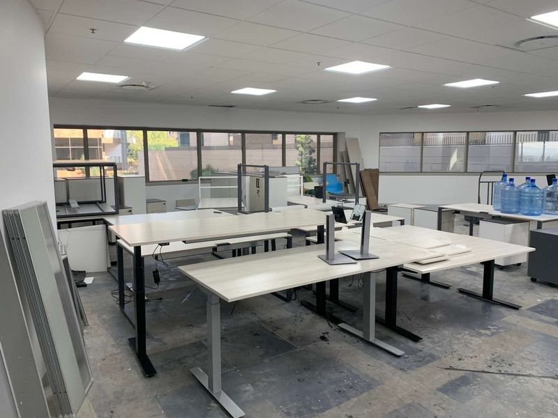 Open-plan office space featuring large windows, suspended ceiling with recessed lighting, and worn flooring, currently set up with multiple workstations.