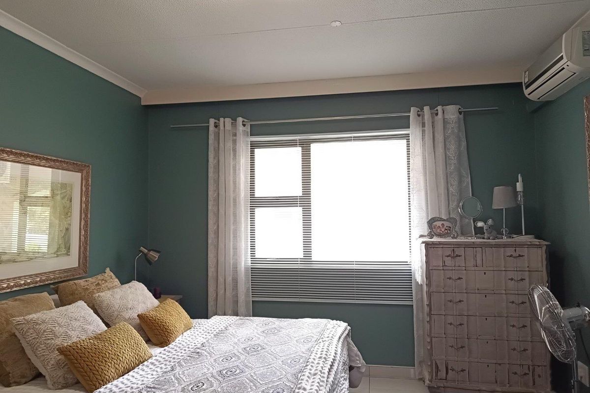 A well-maintained bedroom featuring a wall-mounted air conditioning unit, tiled flooring, and a large window with fitted blinds.