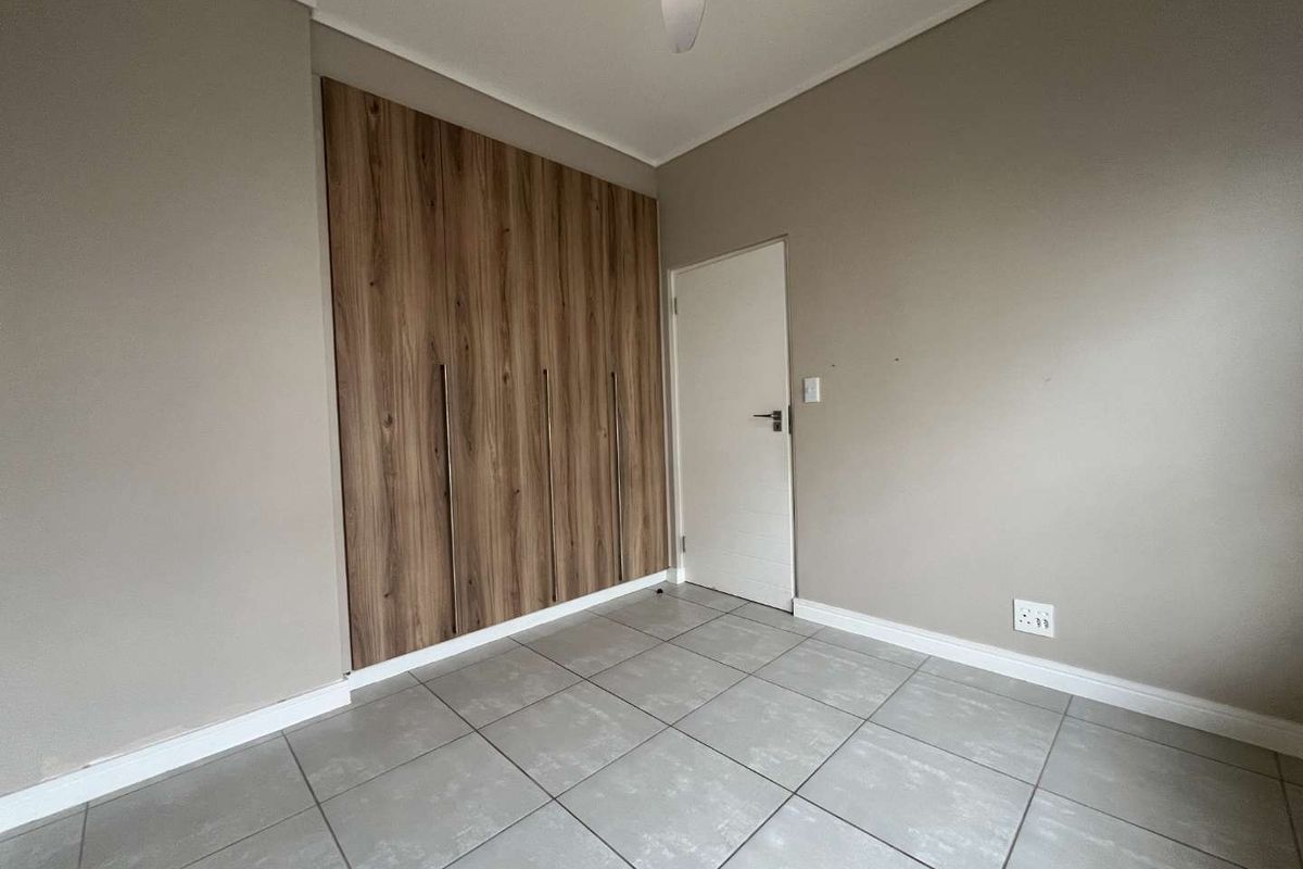 A clean, empty bedroom featuring modern wood-finish built-in cupboards, neutral wall paint, and large format grey floor tiles.