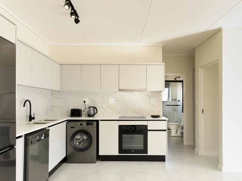 Modern kitchen featuring sleek white cabinetry, marble-look splashback, integrated oven, electric cooktop, and high-quality finishes with an adjacent view into the bathroom.