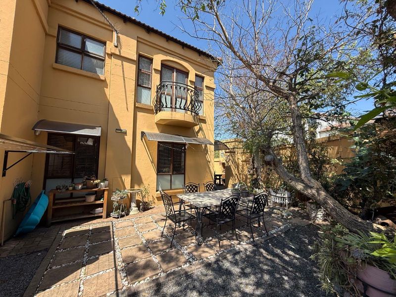 Rear exterior view of the townhouse featuring a paved patio area, a small wrought iron balcony, and established garden trees.