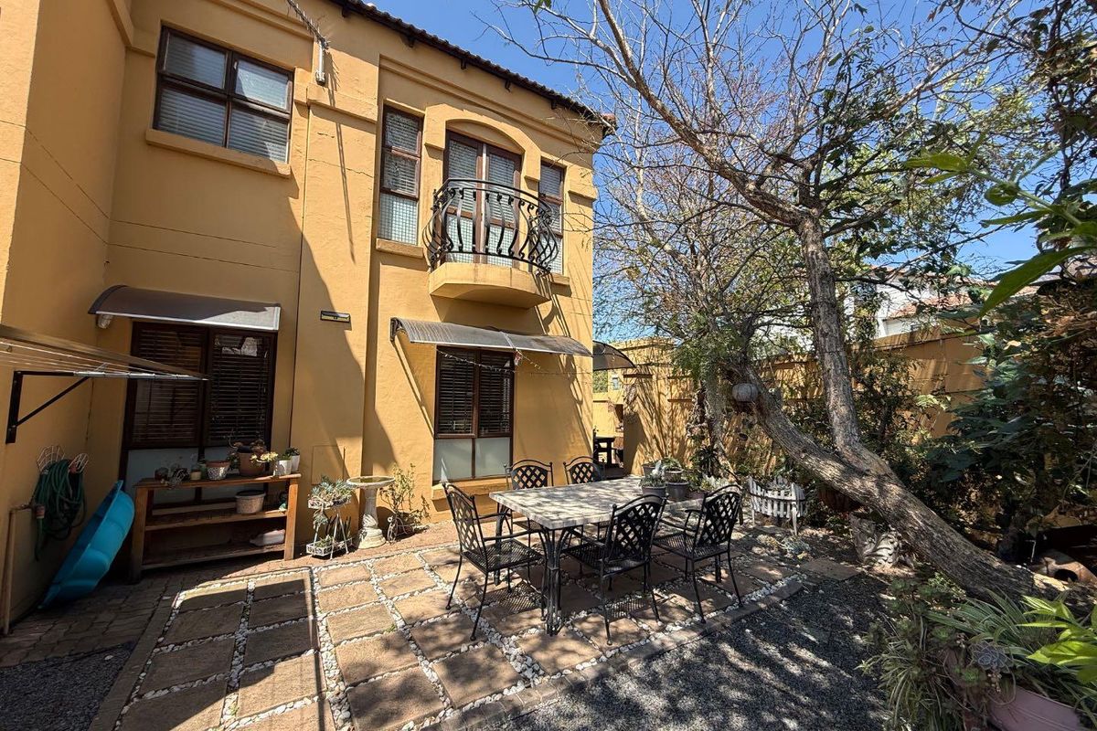 Rear exterior view of the townhouse featuring a paved patio area, a small wrought iron balcony, and established garden trees.