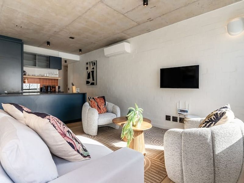 Modern open-plan living area featuring an exposed concrete ceiling, timber flooring, and a sleek kitchen with dark cabinetry and a tiled backsplash.