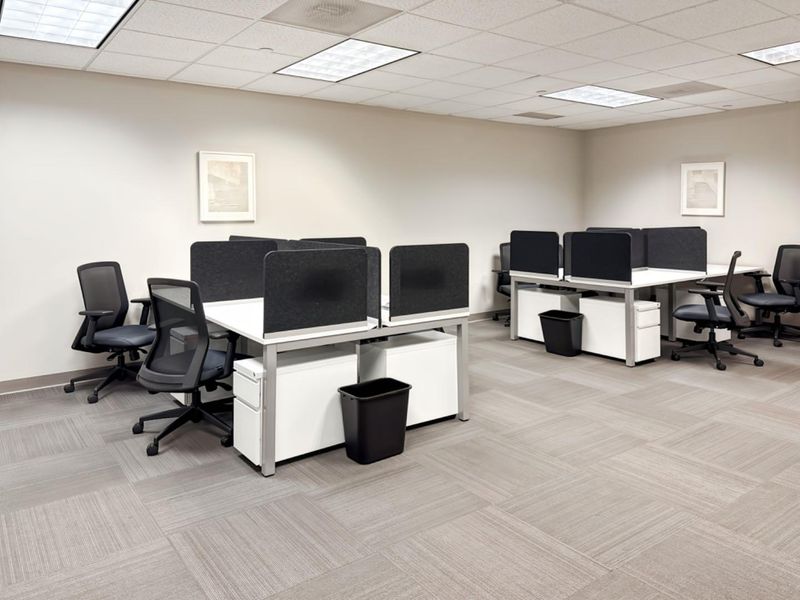 A professional office space featuring modular workstations, suspended ceiling tiles, recessed lighting, and neutral carpet flooring.