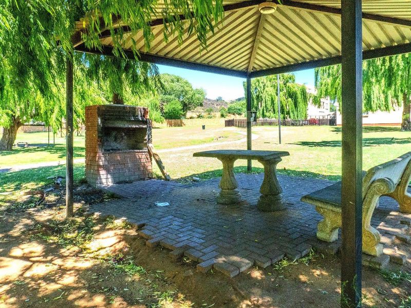 Outdoor covered patio area featuring a built-in brick braai, stone table, and bench seating, set within a communal garden space.