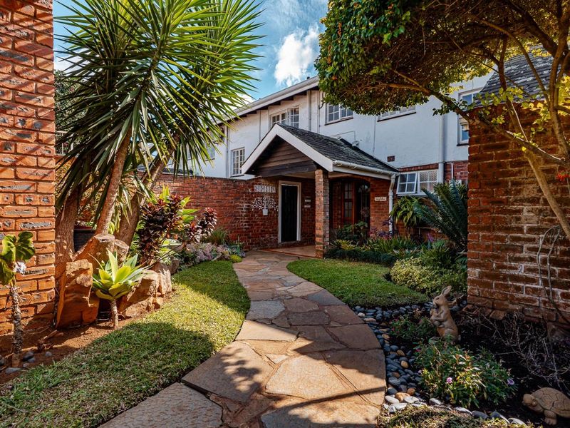 A charming townhouse entrance featuring a stone paved walkway, lush landscaped garden, and a covered porch with classic brick architecture.