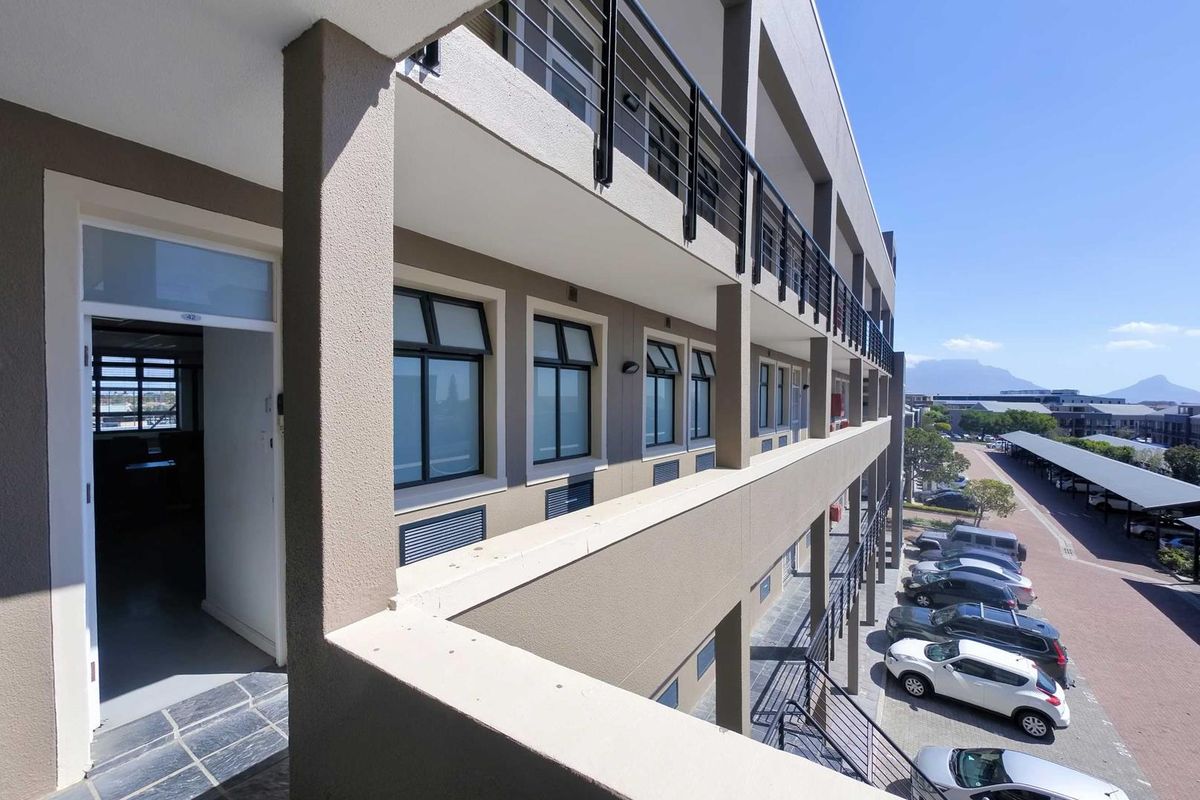 Exterior view of a multi-story office building featuring a covered balcony walkway, metal railings, and paved parking area below.