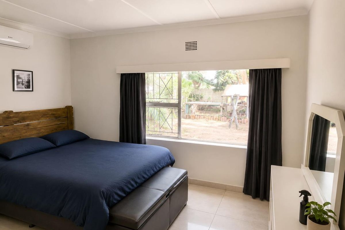 A well-maintained bedroom featuring tiled flooring, an air conditioning unit, and a large window with security bars overlooking the rear garden.