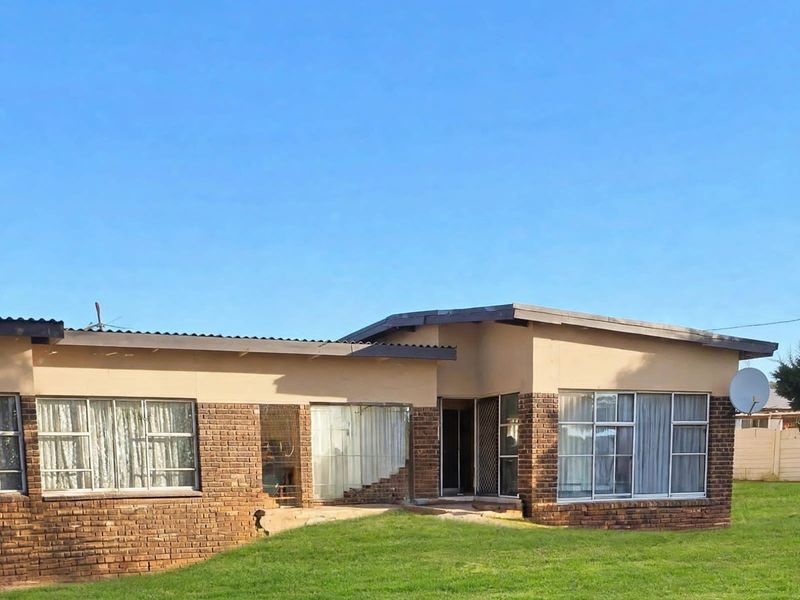Rear view of a single-story brick house featuring large windows, a corrugated metal roof, and a spacious lawn area.