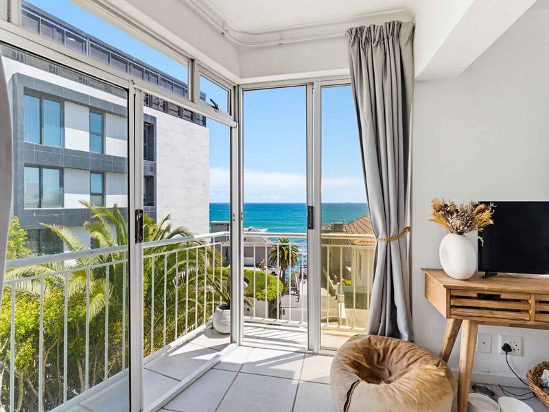 A bright balcony featuring tiled flooring, metal balustrades, and sliding glass doors, offering a clear view of the ocean and surrounding neighborhood.