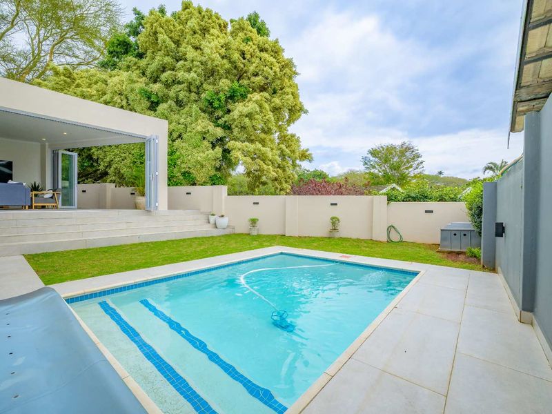 A well-maintained outdoor pool area featuring a paved patio, manicured lawn, and seamless indoor-outdoor flow through large bi-fold doors.