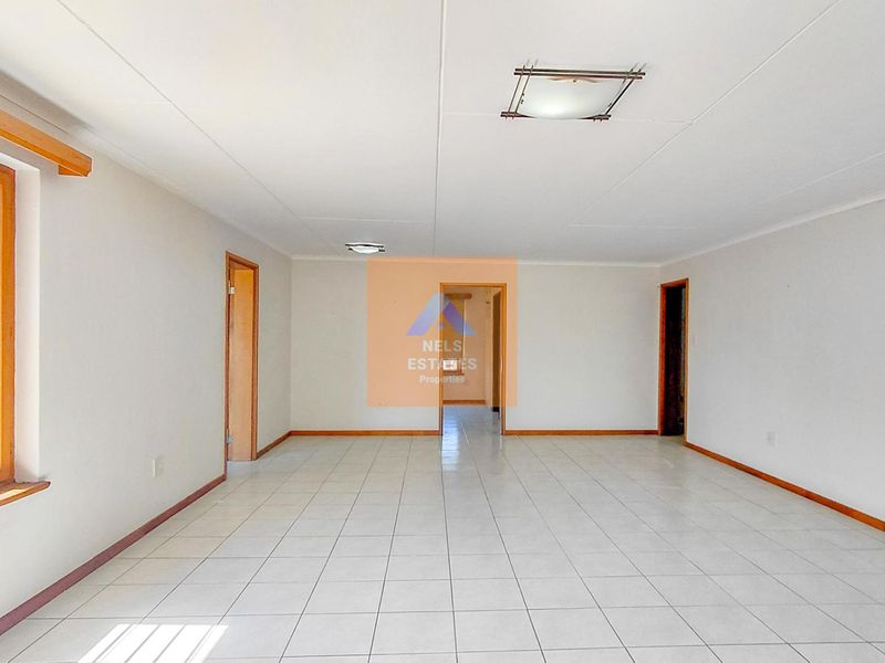 A spacious, empty living area featuring neutral tiled flooring, wooden window frames, and simple ceiling lighting, offering a blank canvas for interior design.