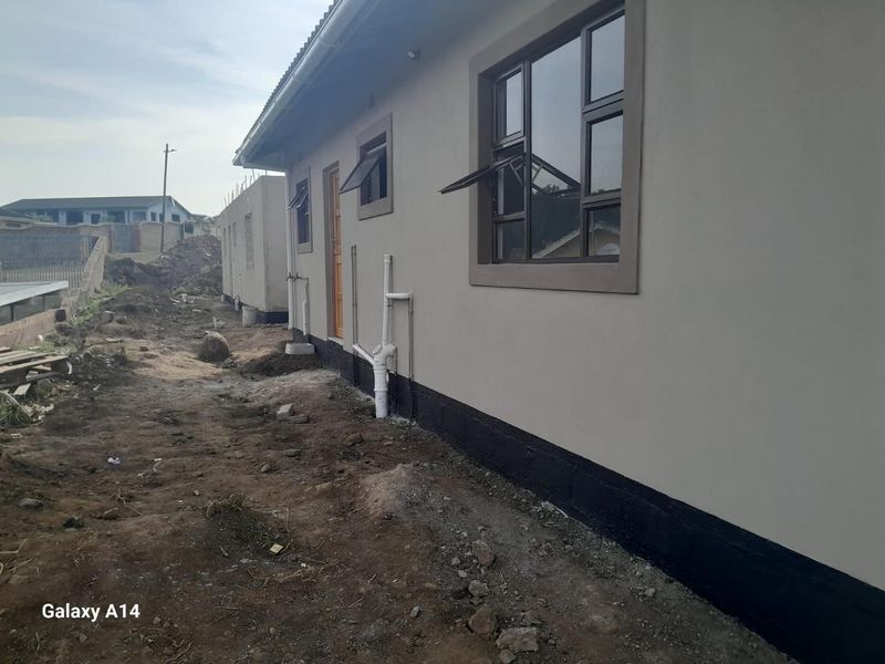 Rear exterior view of a house showing plastered walls, aluminium window frames, and exposed plumbing infrastructure with unfinished surrounding ground.