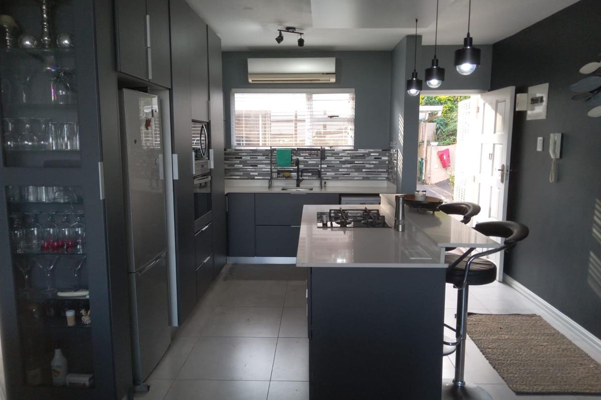 Modern kitchen featuring dark cabinetry, a central island with a gas cooktop, tiled backsplash, and integrated appliances, leading out to an exterior area.