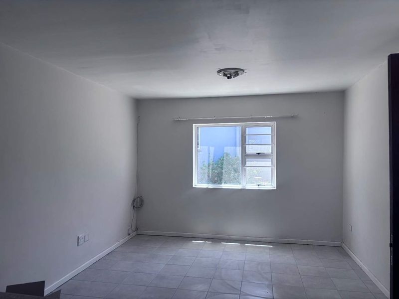 A clean, empty bedroom featuring neutral tiled flooring, a single aluminium window with a curtain rail, and basic electrical fixtures.