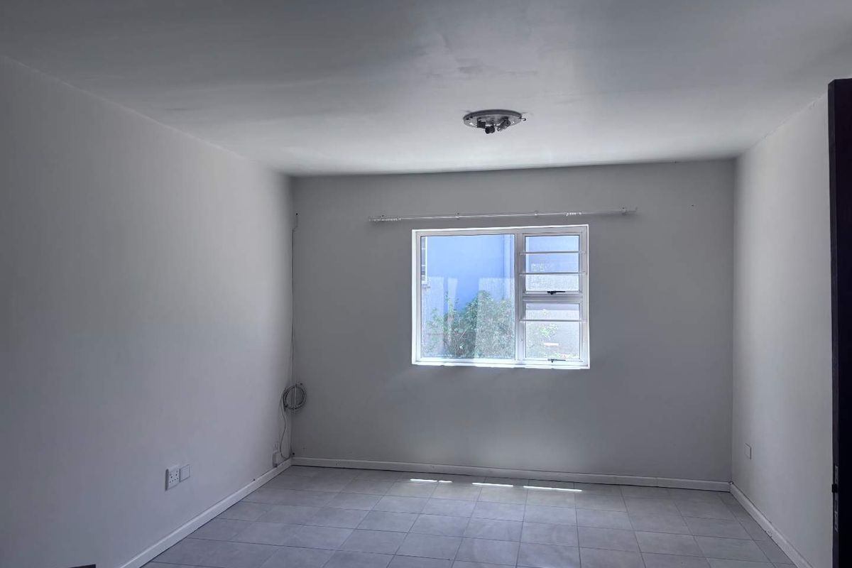 A clean, empty bedroom featuring neutral tiled flooring, a single aluminium window with a curtain rail, and basic electrical fixtures.