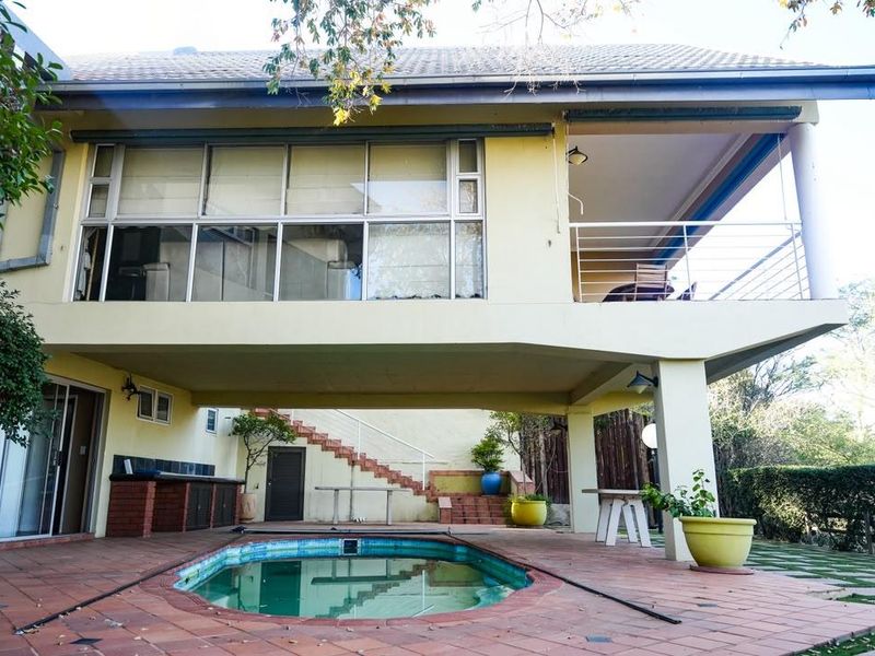 Rear exterior view featuring a swimming pool, paved patio area, built-in brick braai, and an elevated balcony overlooking the garden.