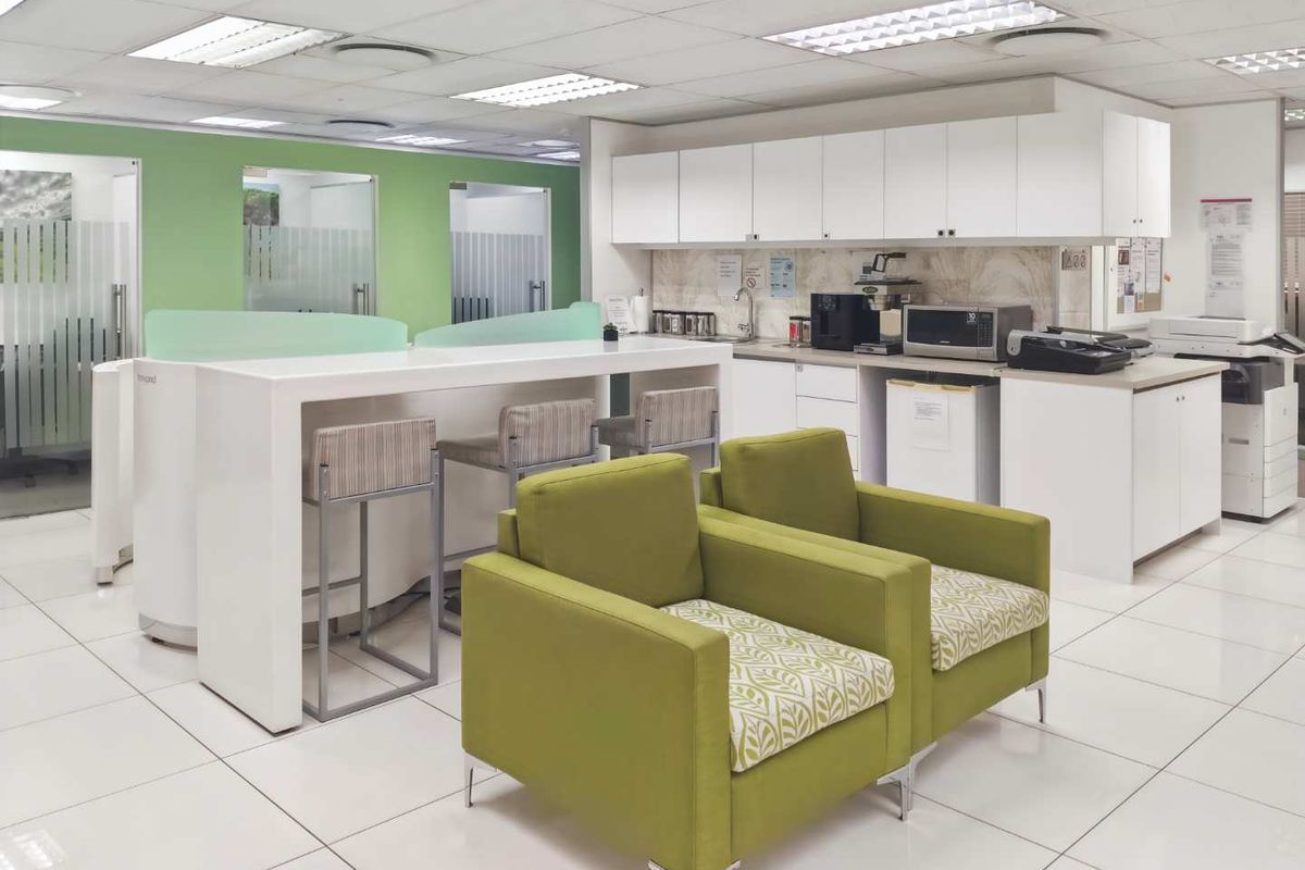 A modern office breakroom featuring a kitchenette with white cabinetry, a breakfast bar, and integrated lighting within a suspended ceiling.