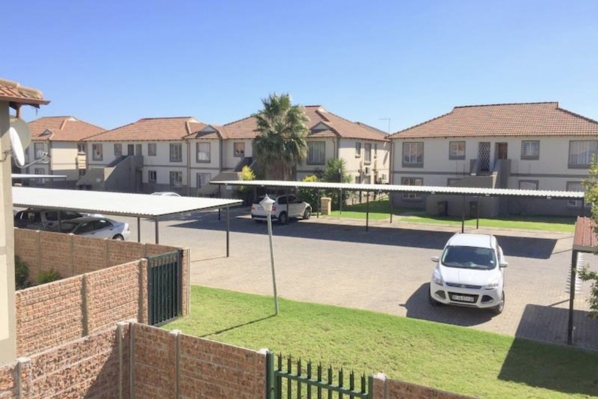 Exterior view of the apartment complex featuring paved parking areas with covered carports, brick boundary walls, and well-maintained communal garden spaces.