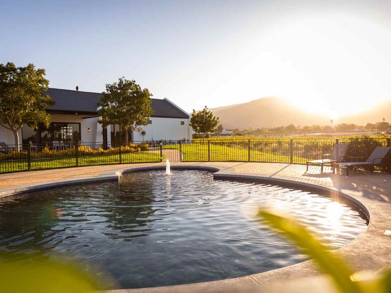 A beautifully maintained swimming pool area featuring a central water fountain, paved decking, and metal perimeter fencing, set against a scenic landscape.