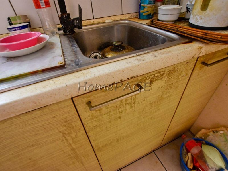 A kitchen sink area featuring laminate cabinetry and a stone-look countertop, showing significant wear and deterioration on the cabinet surfaces and faucet fixtures.