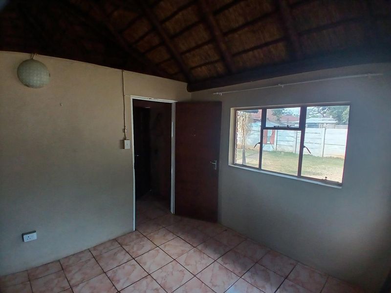 An empty bedroom featuring a traditional thatched roof with exposed timber rafters, tiled flooring, and a metal-framed window overlooking the garden.