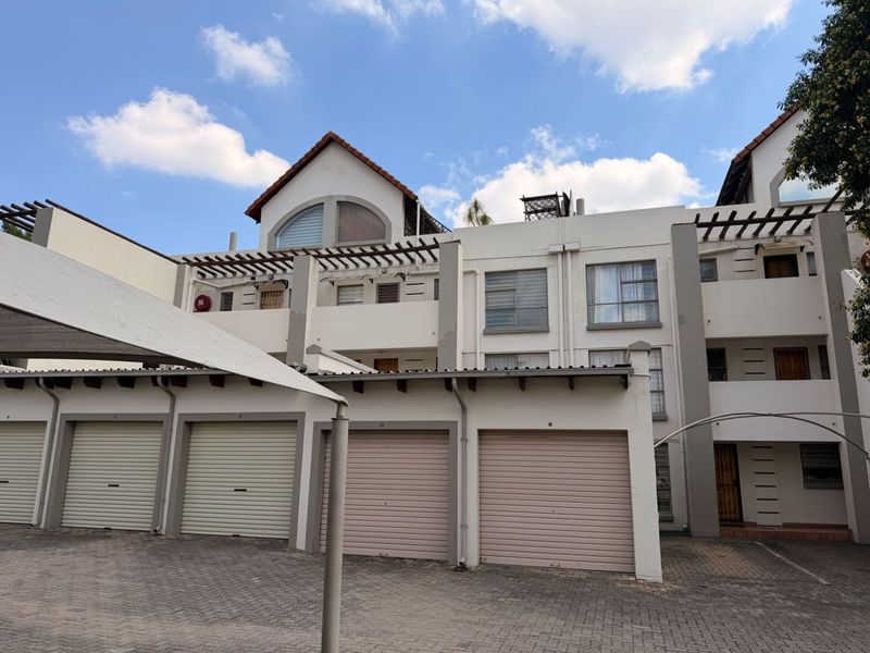 Front elevation of a multi-story apartment complex featuring multiple roller shutter garage doors, paved driveway, and upper-level balconies with pergola structures.