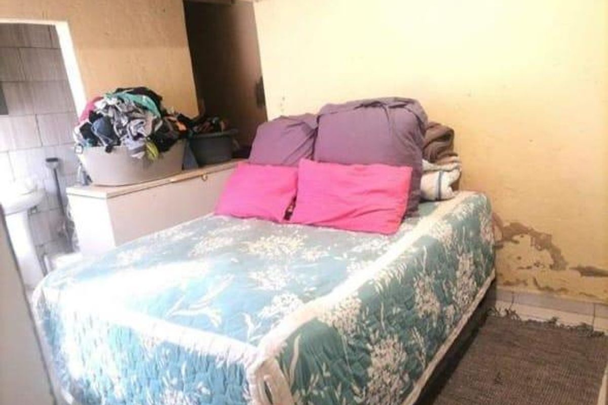 A bedroom featuring a bed with visible wall damage and peeling paint, adjacent to a small utility area with a sink.