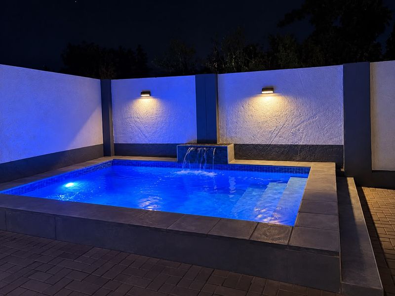 A well-maintained swimming pool area at night, featuring integrated water feature, modern wall lighting, and clean paved surround.