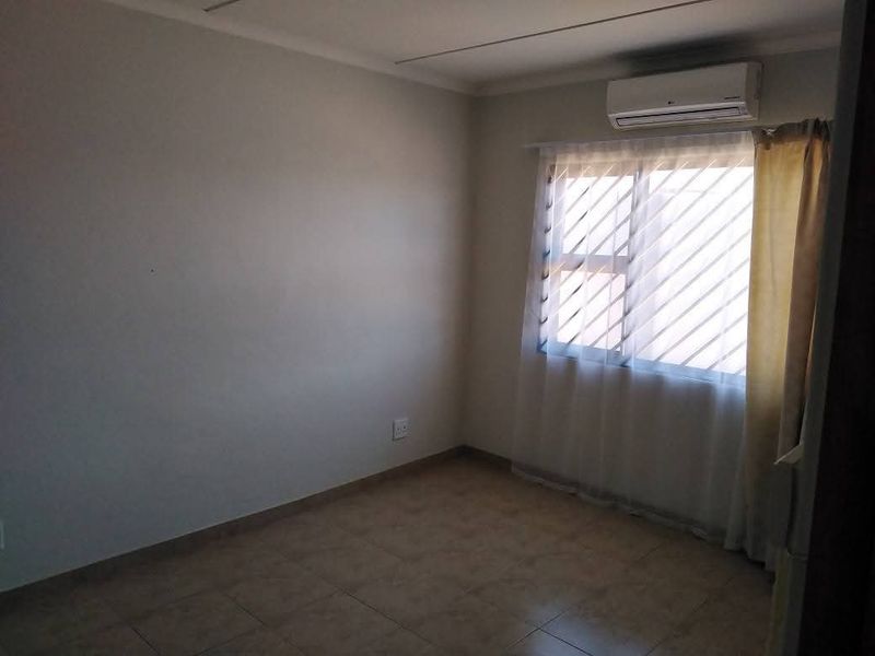 A clean, empty bedroom featuring tiled flooring, a louvered window with security bars, and a wall-mounted air conditioning unit.