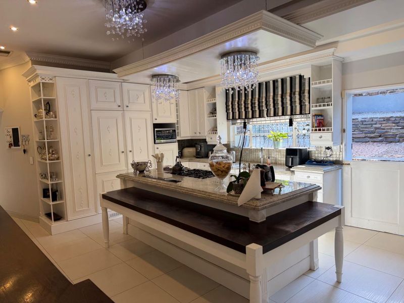 A well-appointed kitchen featuring custom white cabinetry, granite countertops, a central island with a gas hob, and elegant crystal chandelier lighting.