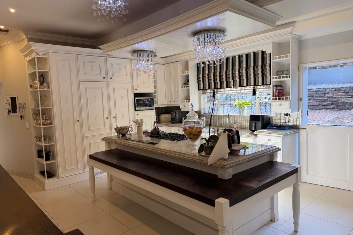 A well-appointed kitchen featuring custom white cabinetry, granite countertops, a central island with a gas hob, and elegant crystal chandelier lighting.