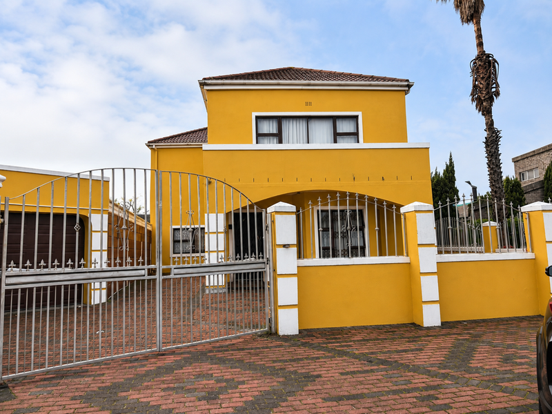 Front elevation of a two-story house featuring a yellow facade, metal security gate, and a brick-paved driveway.