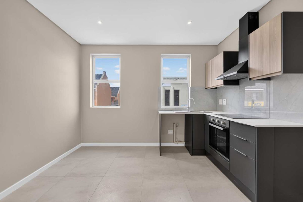 Modern, empty kitchen featuring sleek dark grey and wood-tone cabinetry, stone countertops, integrated oven, hob, and extractor fan with neutral tiled flooring.