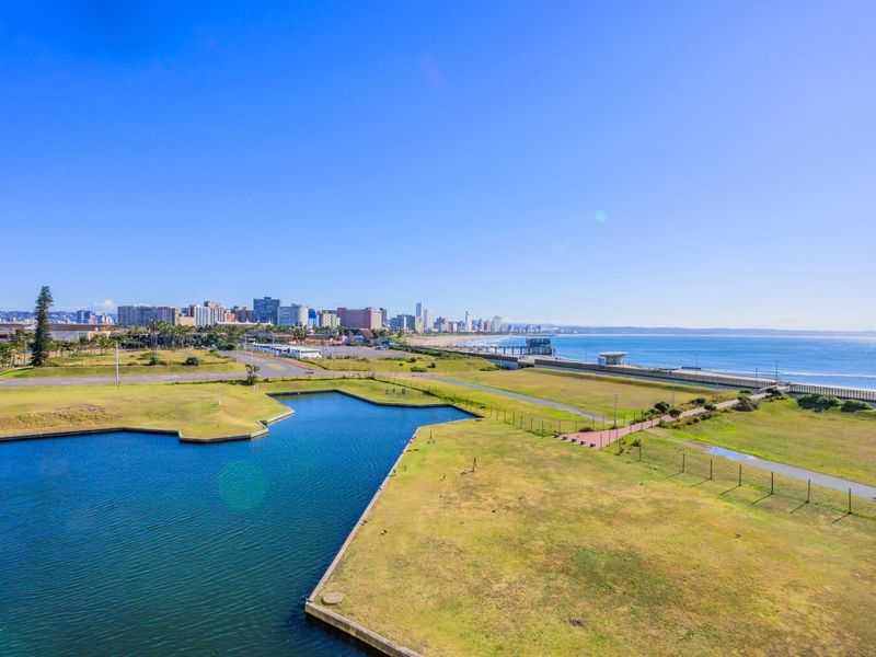 Panoramic coastal view featuring a landscaped park, a small water body, and the city skyline along the coastline.
