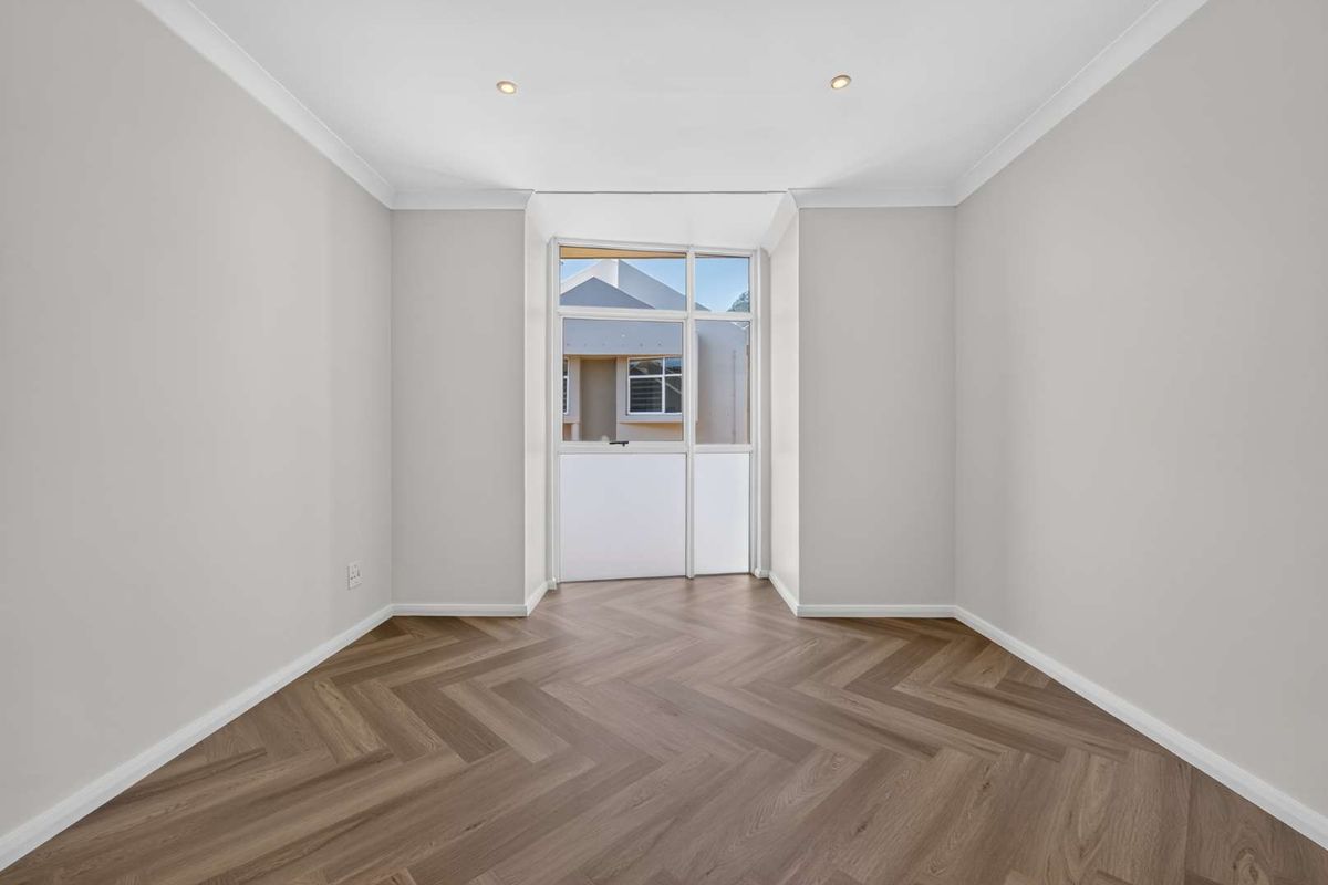 A bright, empty bedroom featuring modern herringbone vinyl flooring, recessed downlighting, and neutral wall finishes.