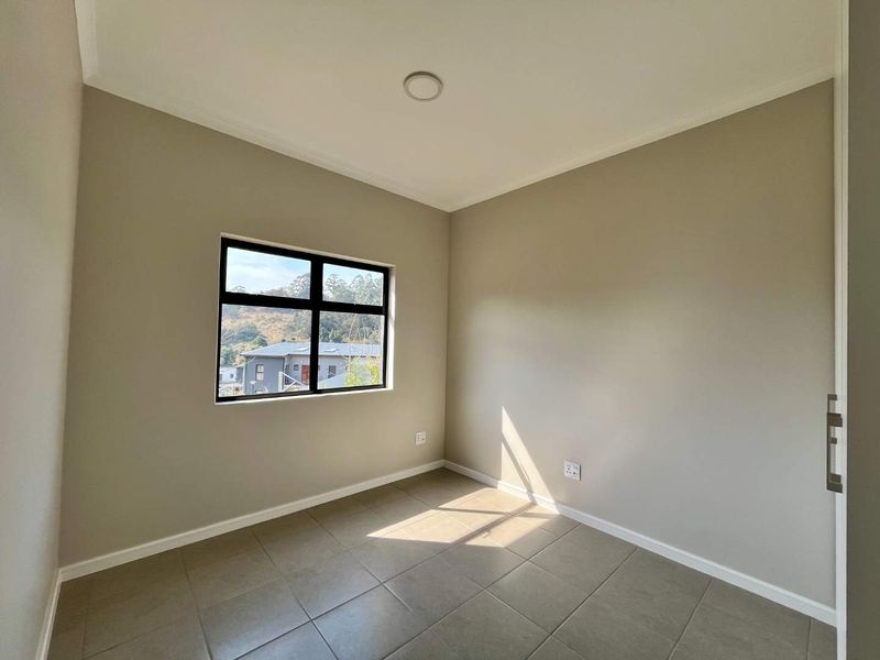 A bright, empty bedroom featuring neutral walls, tiled flooring, and a black aluminium window frame with a view of the surrounding area.