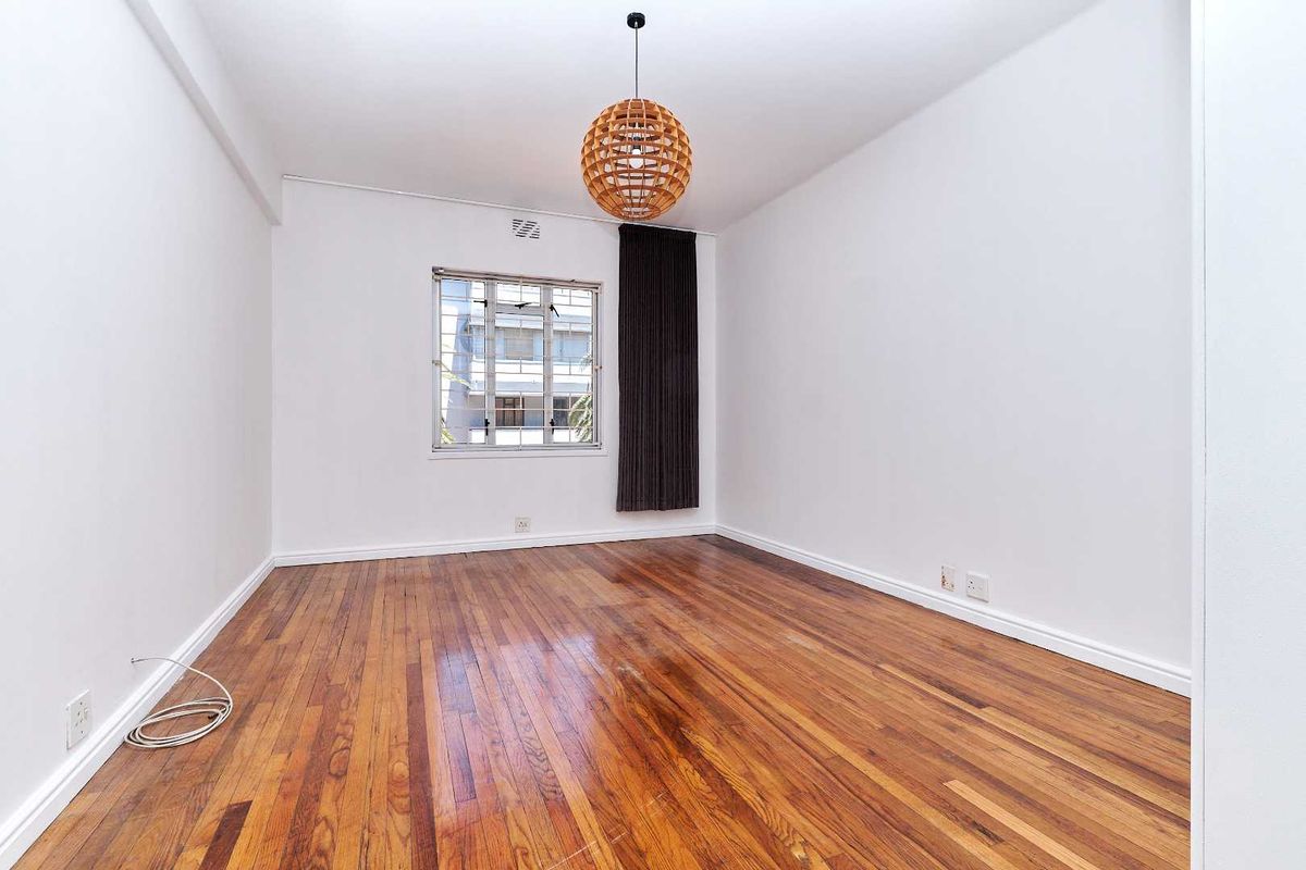 A bright, empty bedroom featuring polished hardwood flooring, white walls, and a single casement window.