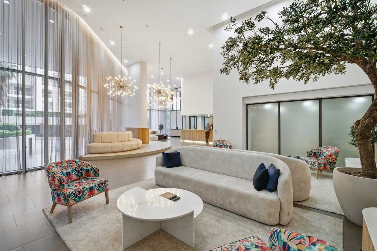 Spacious modern lobby area featuring high ceilings, floor-to-ceiling windows with sheer curtains, recessed lighting, and a contemporary reception desk.