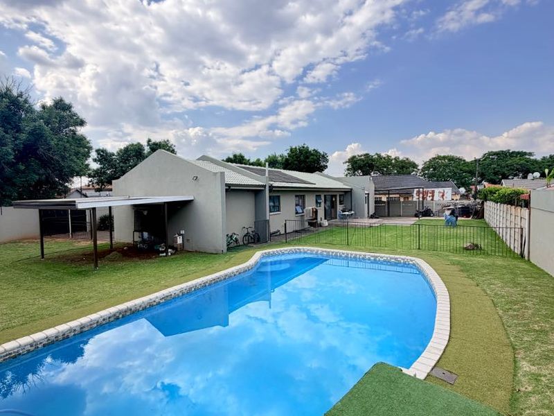 A well-maintained backyard featuring a swimming pool with tiled coping, artificial grass surrounds, metal fencing, and an attached carport structure.