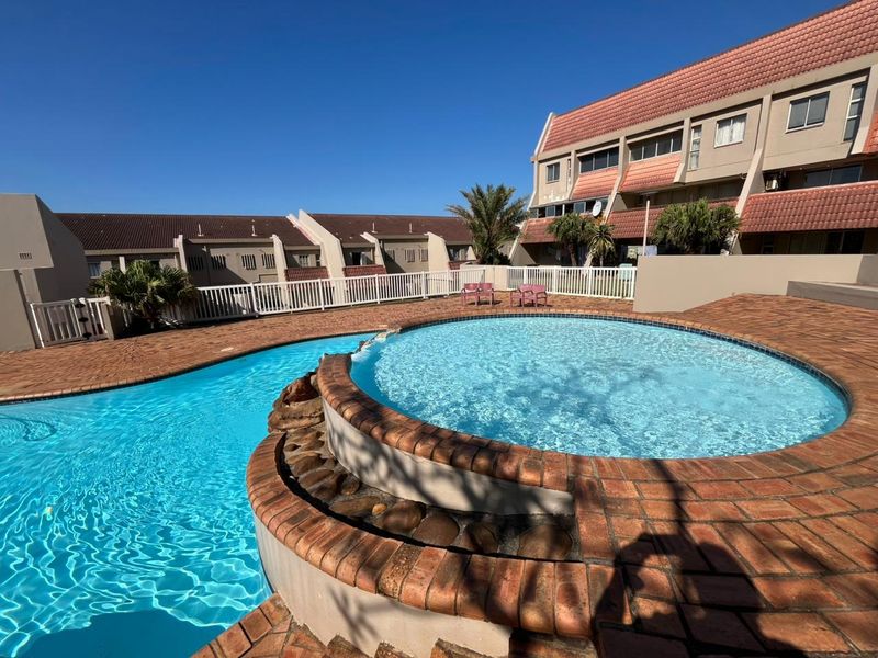 A well-maintained communal swimming pool area featuring brick paving and perimeter fencing, set within an apartment complex with tiled roof architecture.