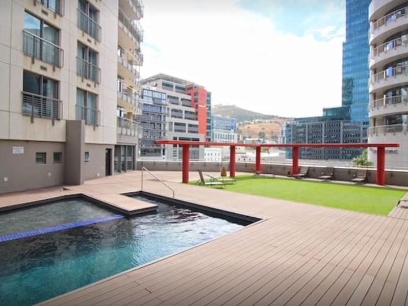 A well-maintained rooftop pool area featuring timber decking, artificial grass, and a modern red steel pergola with views of the surrounding city buildings.