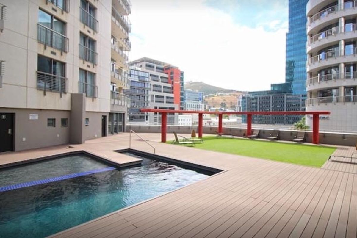 A well-maintained rooftop pool area featuring timber decking, artificial grass, and a modern red steel pergola with views of the surrounding city buildings.